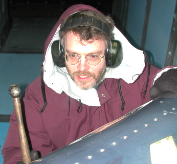 A bearded, rather handsome young man, wearing a cold-weather coat, gloves, and ear protection. He is holding a hammer, ready to strike a test airfoil.